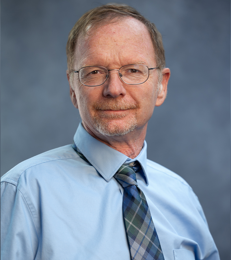 David Rose wearing a blue dress shirt with a patterned tie standing in front of a blueish grey photo backdrop