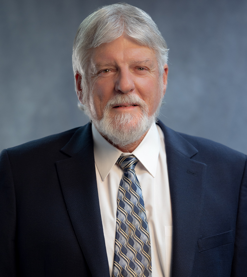 Dr. Gary Thomas Scott wearing a dark blue blazer, a white dress shirt, and a blue with yellow tie standing in front of a blueish grey photo background