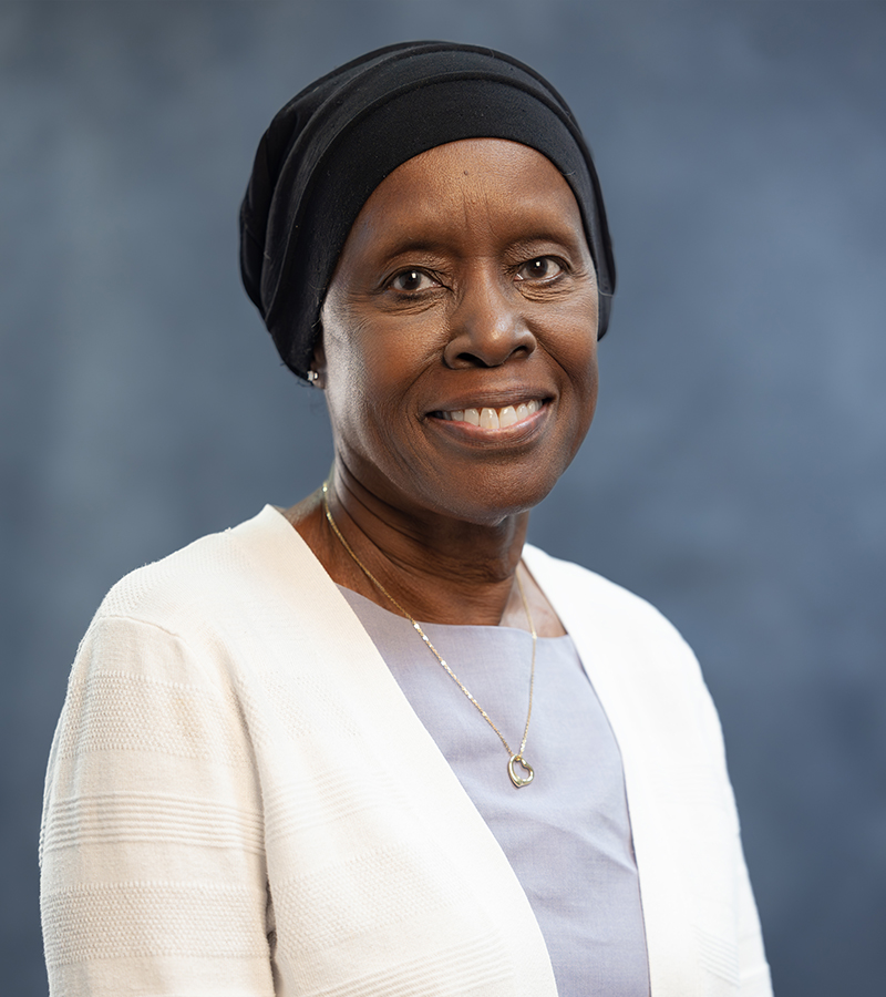 Helen Hall wearing a white sweather with a purple dress standing in front of a blueish grey photo backdrop
