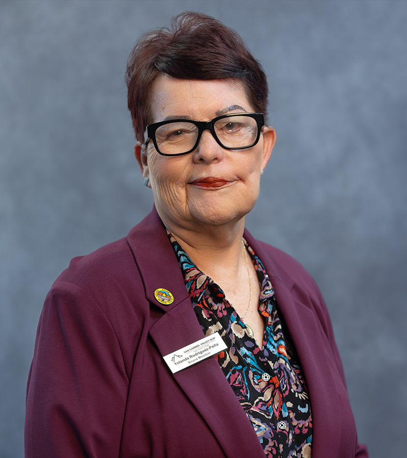 Yolanda Rodriguez-Peña wearing a maroon blazer with a pattern blue and red top standing in front of a blueish grey photo backdrop