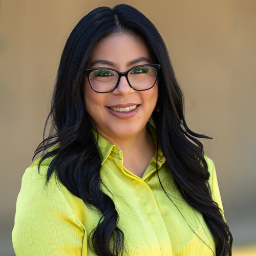 Professional headshot of a person wearing glasses and a bright yellow blouse, smiling at the camera against a softly blurred background.