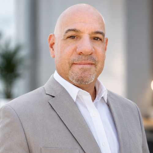 Professional headshot of a person in a light gray blazer and white dress shirt, standing indoors against a softly blurred background and looking at the camera.